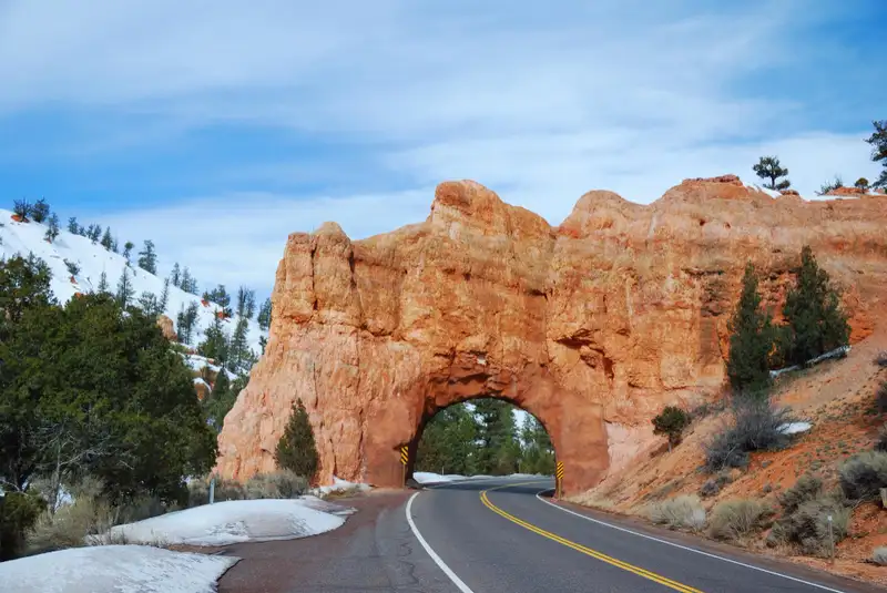A natural arch of reddish-orange rock frames a paved road in a winter landscape. The arch is carved into a large rock formation that spans the width of the road, creating a tunnel-like passage. Patches of snow are visible on the ground to the left of the road and further up the snow-covered slopes on either side of the arch. Pine trees, some dusted with snow, are scattered across the landscape. The sky above is a mix of blue and white clouds, suggesting a partly cloudy winter day. The road has a double yellow line marking the center.